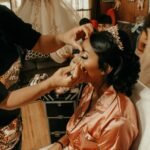 A bride in peach robe getting her makeup done by an artist before her wedding celebration.